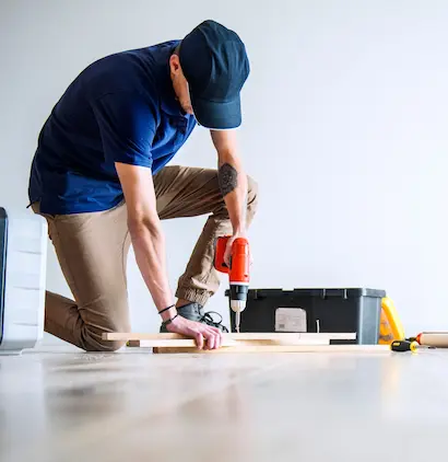 Mężczyzna w niebieskiej koszulce i czapce używa wiertarki do pracy z drewnem, klęcząc na jednej nodze. W tle widoczna jest skrzynka narzędziowa.